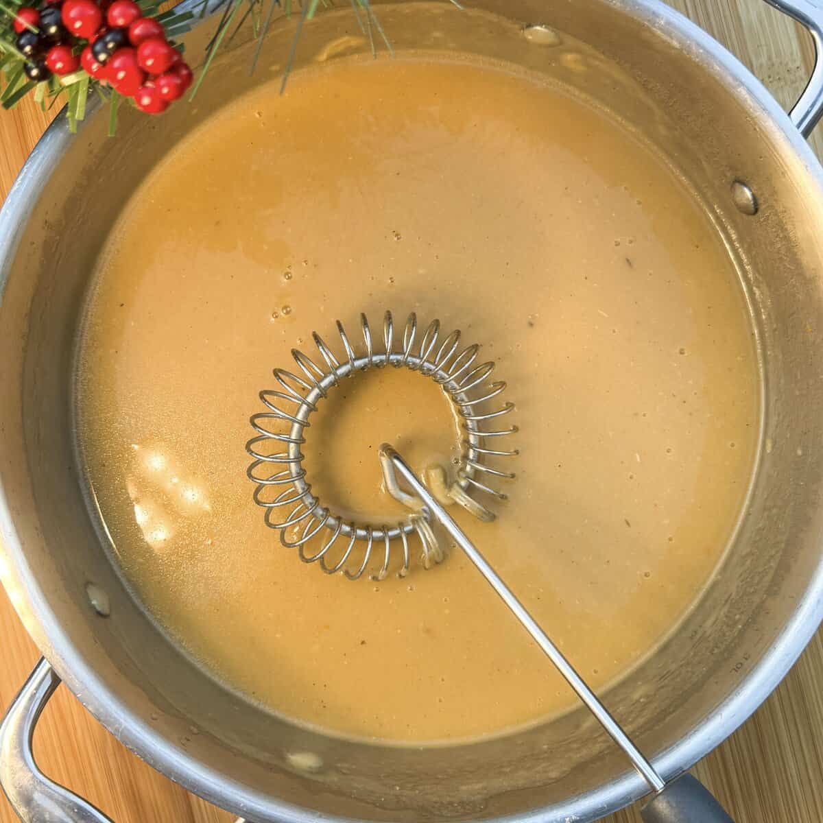 A pot of creamy brown gravy being whisked, with a festive sprig of red berries and pine needles visible at the top left corner. The pot sits on a wooden surface.