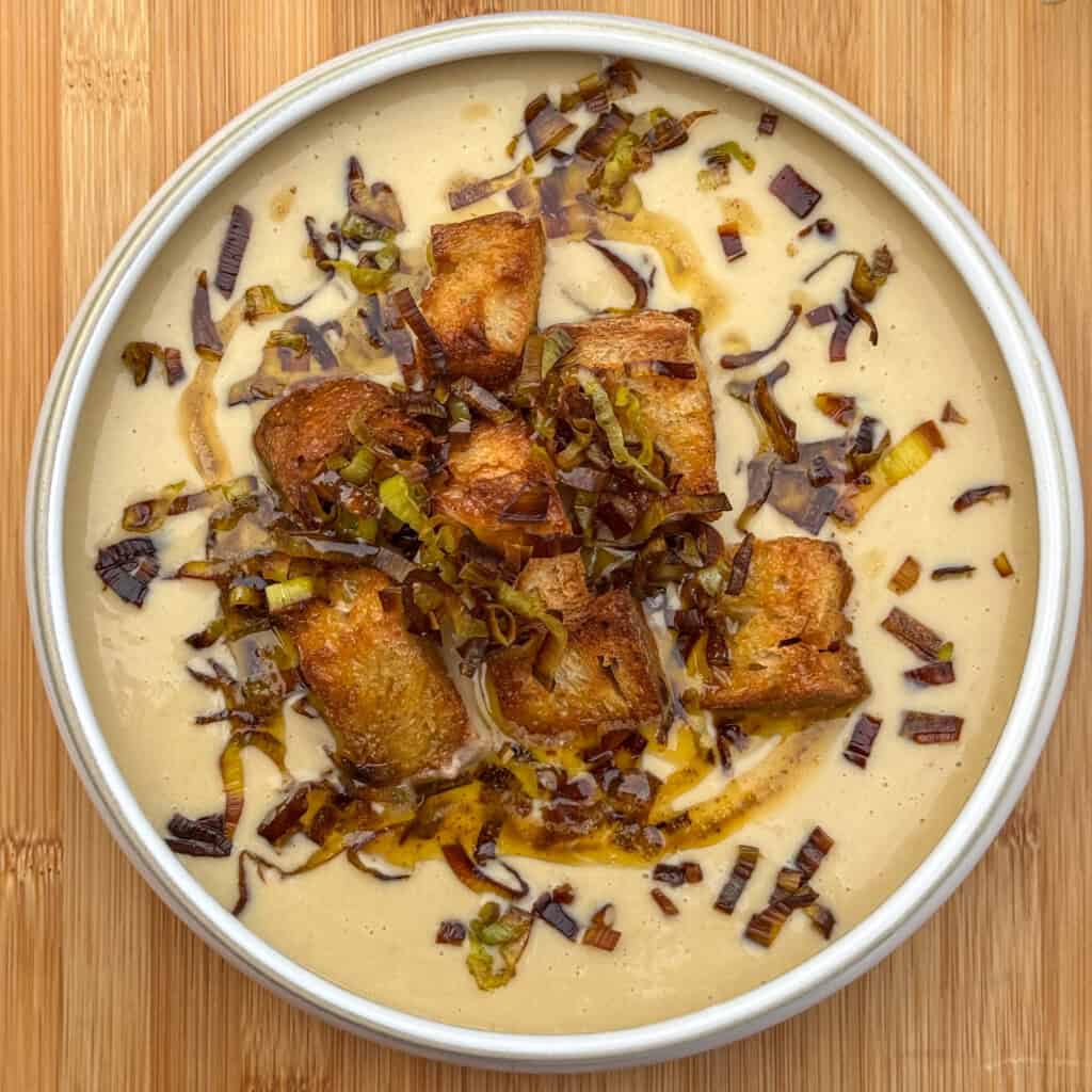 A bowl of creamy leek and potato soup topped with golden croutons and sautéed leeks, set on a wooden surface. The soup is smooth and light in color, with a drizzle of oil and scattered crispy toppings.