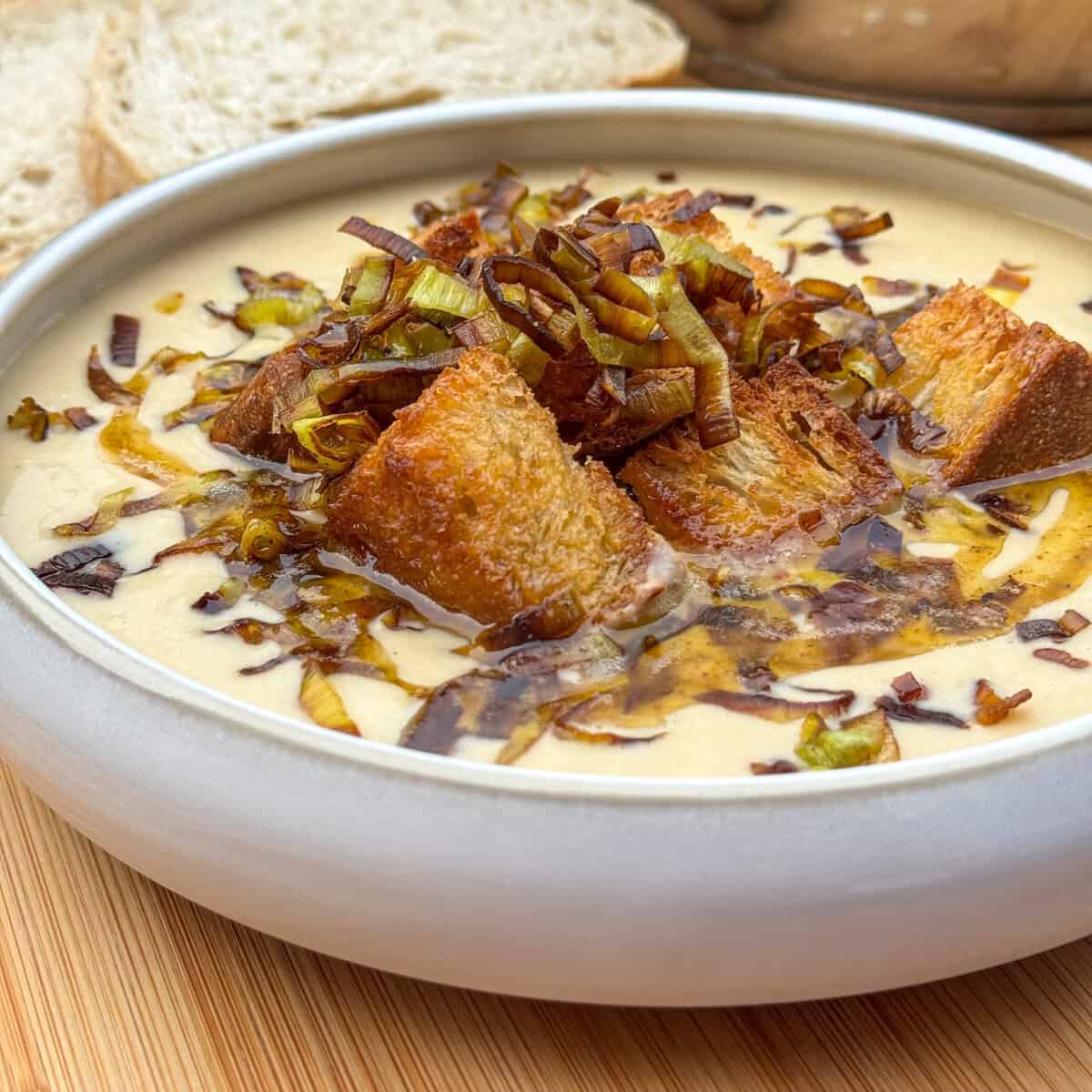 A bowl of creamy leek and potato soup topped with golden brown croutons, sautéed leeks, and a drizzle of oil, with slices of bread in the background on a wooden surface.