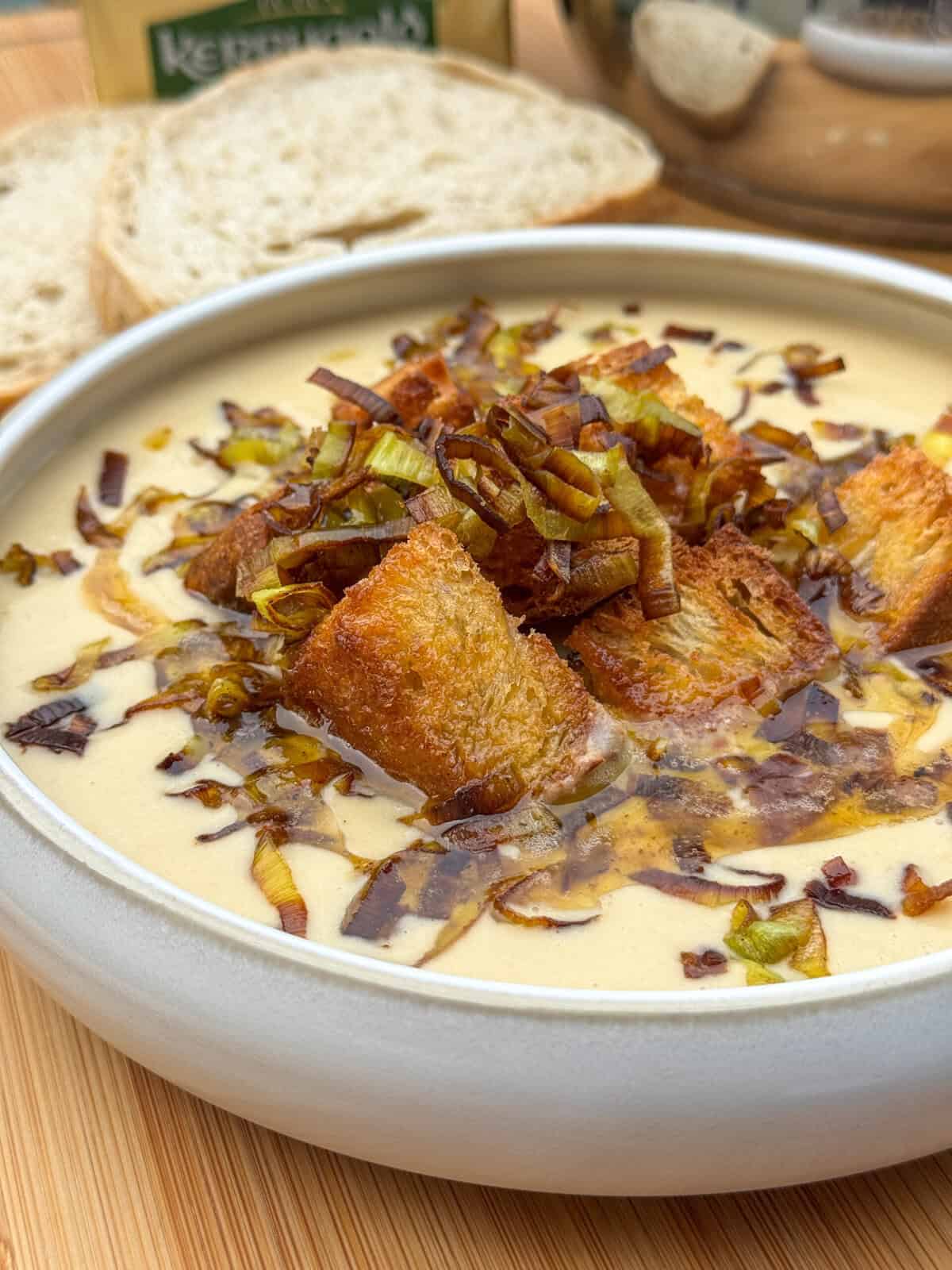 A bowl of creamy potato and leek soup topped with golden croutons and sautéed leeks, served with slices of rustic bread in the background on a wooden surface.