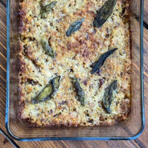 A glass baking dish filled with golden-brown stuffing, topped with browned, crispy sage leaves, resting on a wooden surface.
