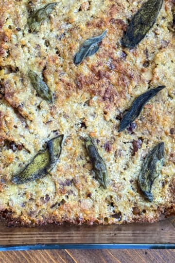 A glass baking dish filled with golden-brown stuffing, topped with browned, crispy sage leaves, resting on a wooden surface.