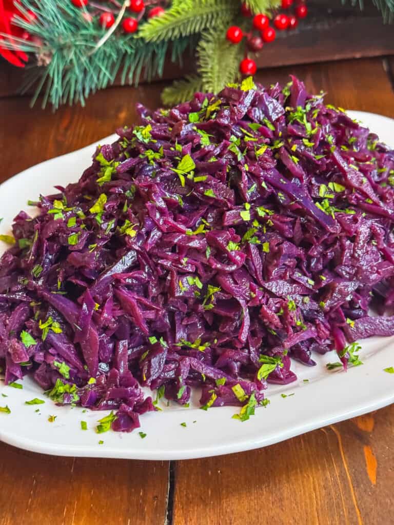 A white plate piled with cooked red cabbage, garnished with chopped fresh parsley. There are pine branches and red berries in the blurred background, suggesting a festive setting.