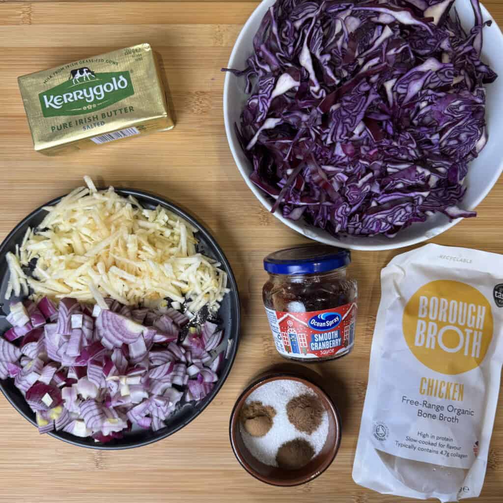 A wooden surface with bowls of shredded red cabbage, grated apple, chopped red onion, a block of Kerrygold butter, a jar of cranberry sauce, a packet of chicken bone broth, and a dish of salt, sugar, and spices.