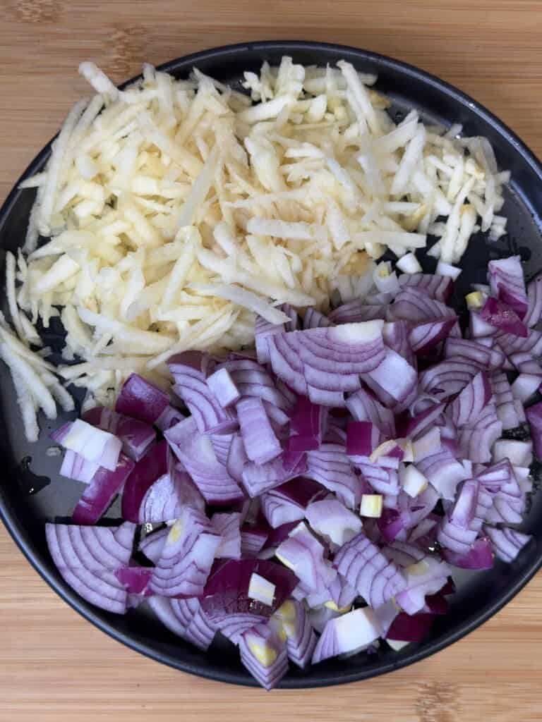 A black plate on a wooden surface holds two piles: finely grated apple on the left and chopped red onion on the right.