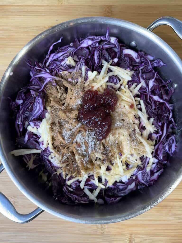A pot filled with shredded red cabbage, topped with grated apple, a spoonful of red jam, and a sprinkle of spices, all ready to be mixed together on a wooden surface.