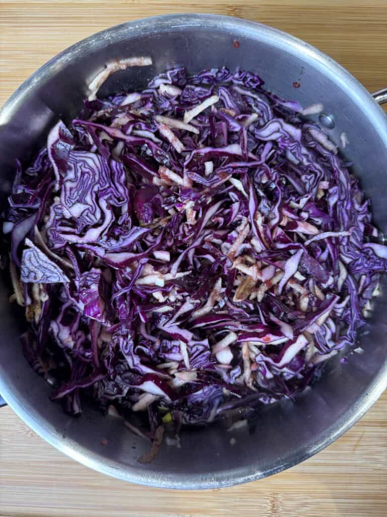 Shredded red cabbage and pieces of apple in a stainless steel pot on a wooden surface, ready to be cooked.