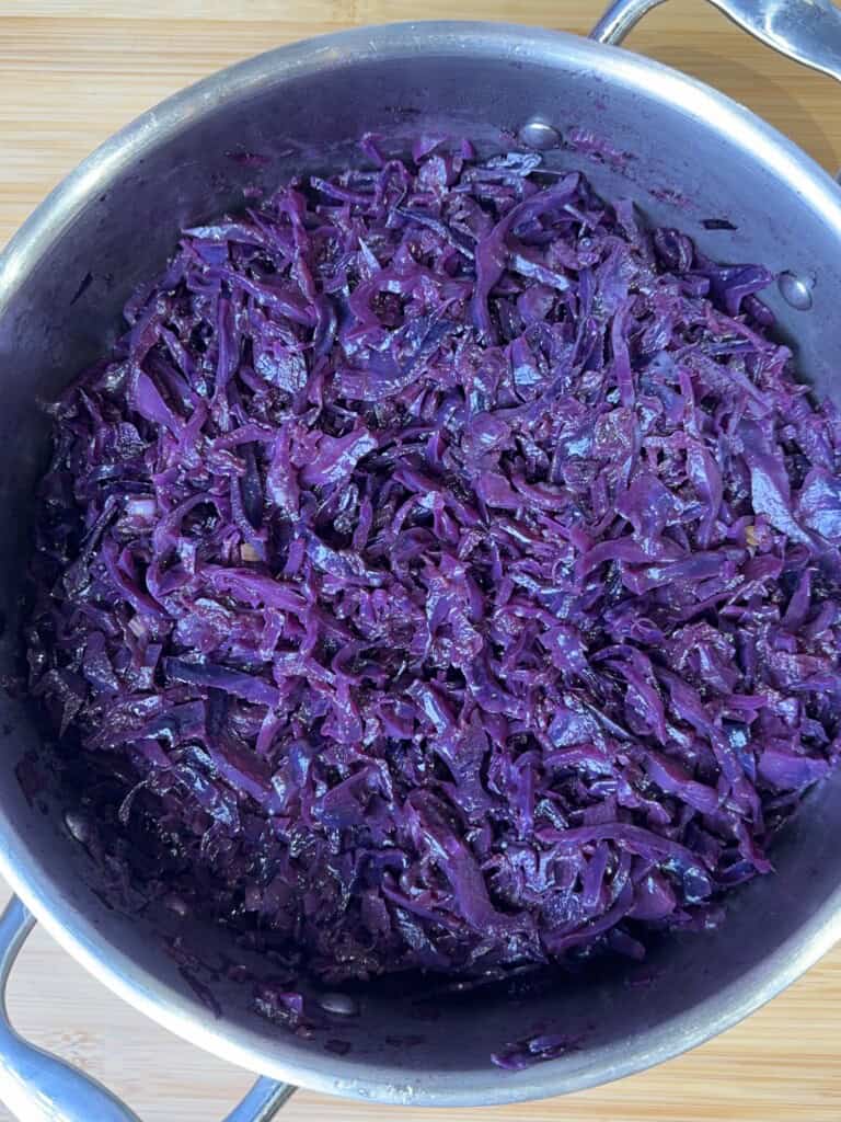 A stainless steel pot filled with cooked, shredded red cabbage sits on a wooden surface. The vibrant purple cabbage appears glossy and tender.