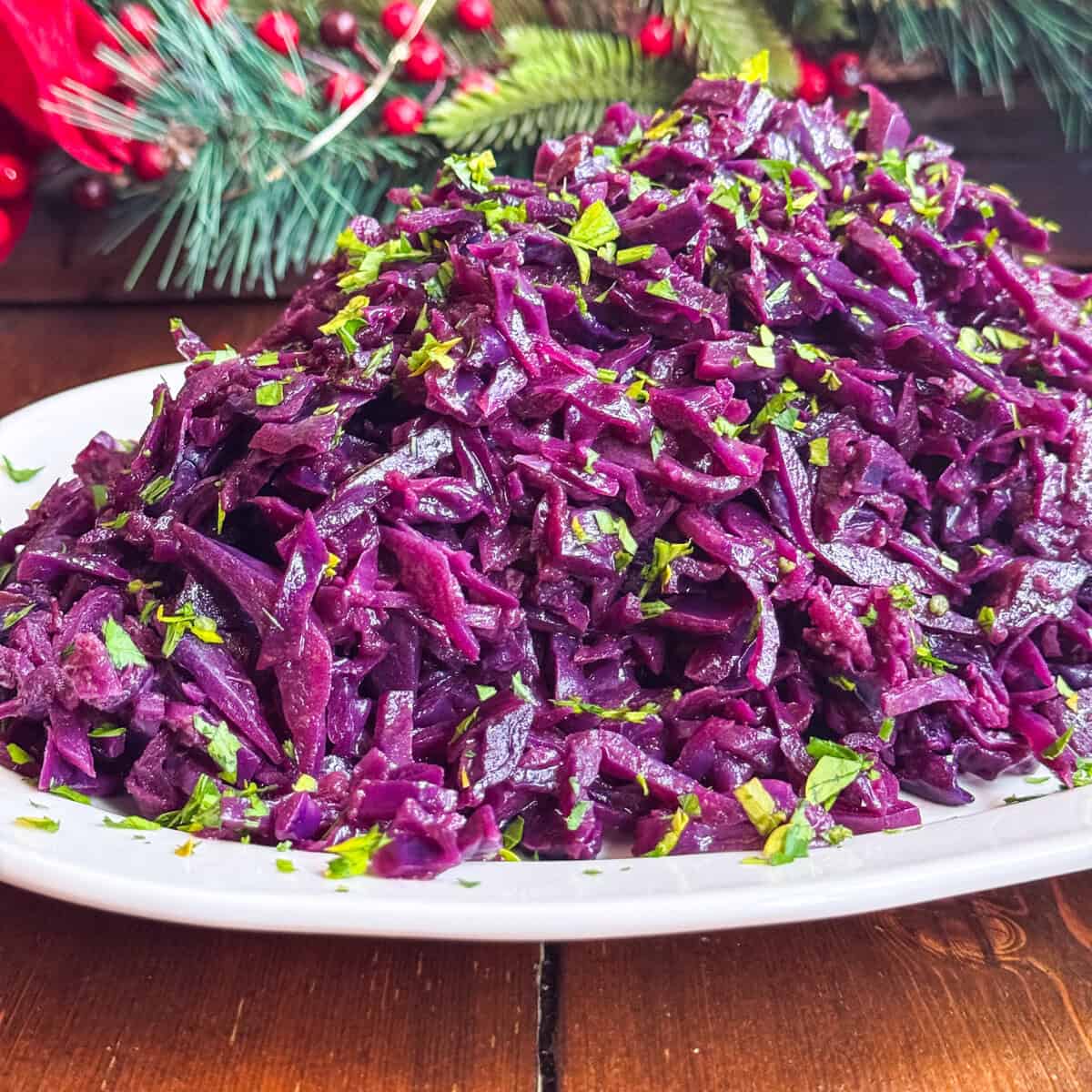 A plate of vibrant spiced red cabbage slaw garnished with chopped herbs, set on a wooden table with pine branches and red berries in the background.