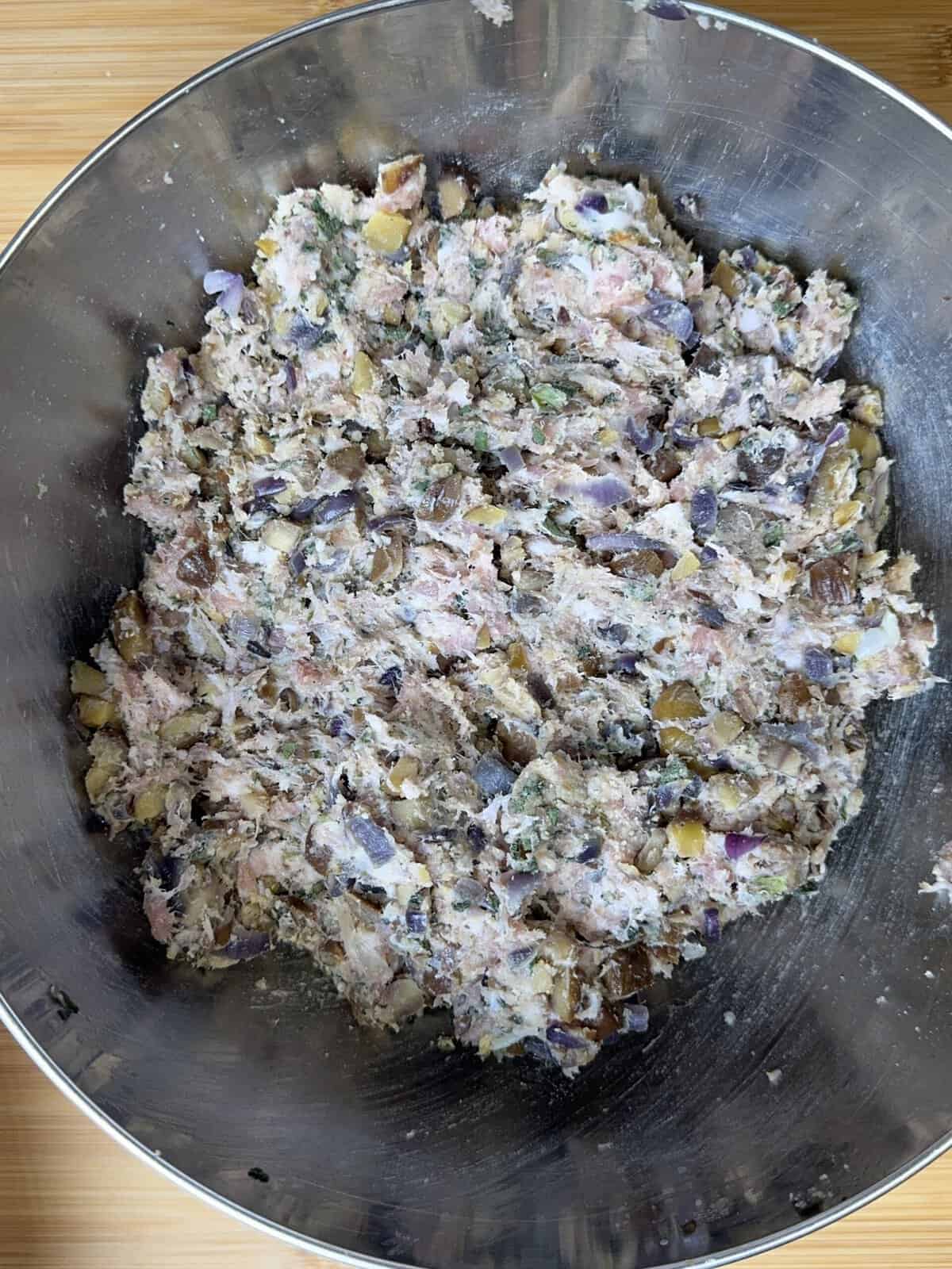 A large metal mixing bowl filled with a sausage meat stuffing mixture of chopped ingredients, including visible purple onions, herbs, and what appears to be fish, ready to be cooked or formed into patties.