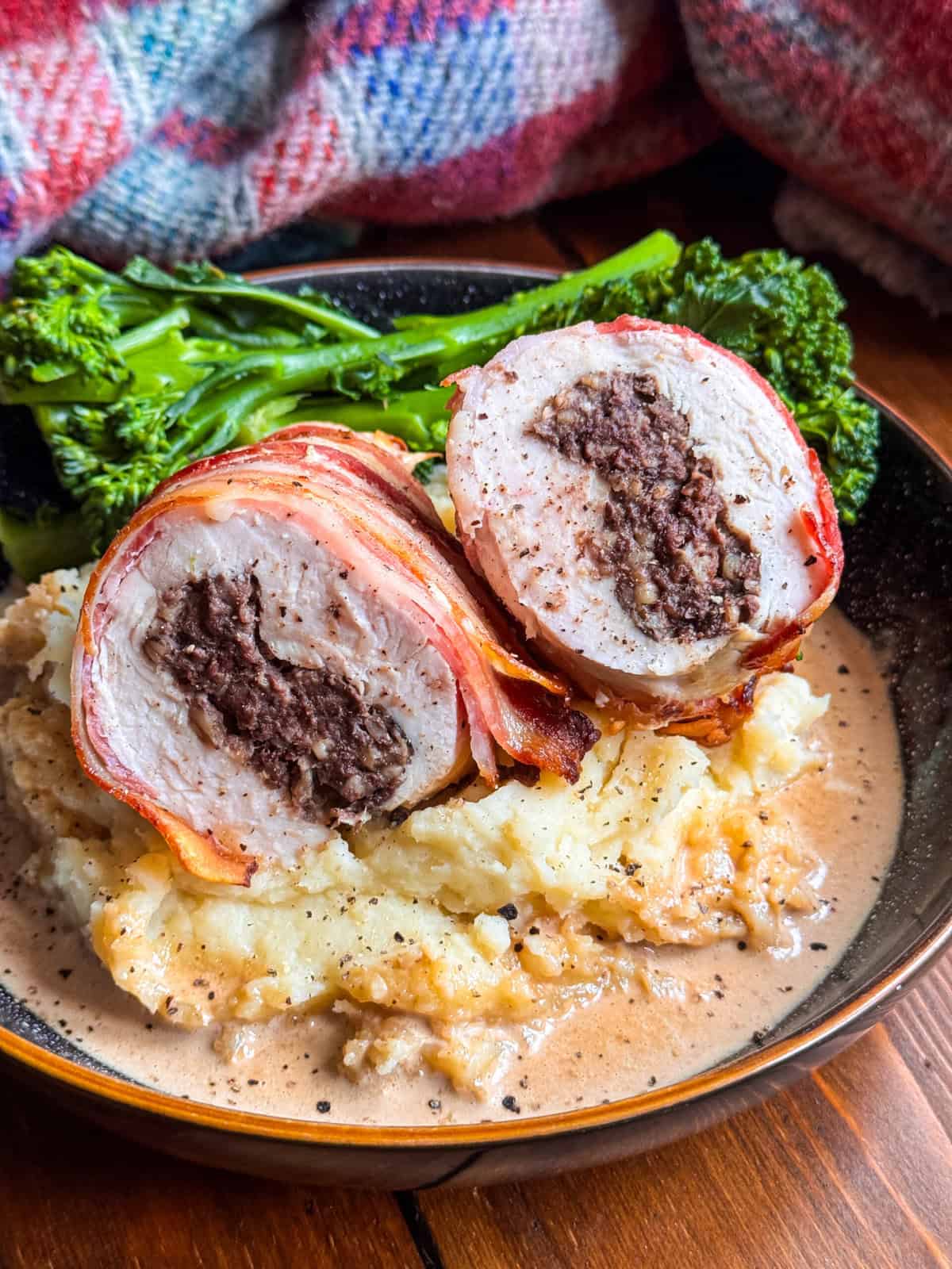 A close-up of Chicken Balmoral, bacon-wrapped haggis stuffed chicken breast, sliced to show a haggis filling, served on mashed potatoes with creamy sauce, and steamed broccolini in the background. A colorful, plaid fabric is partially visible behind the plate.