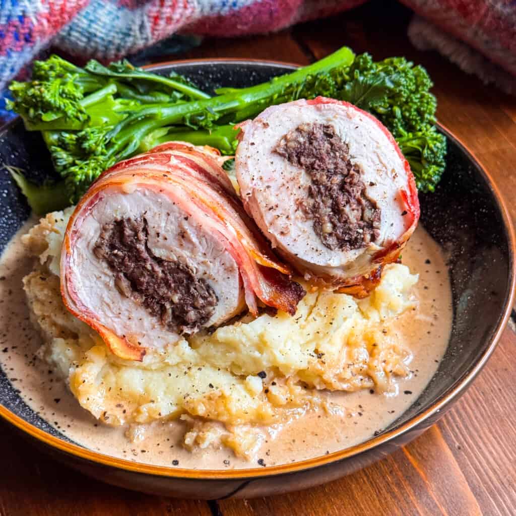 A plate with Chicken Balmoral: bacon-wrapped chicken stuffed with haggis, sliced to show the filling, served on creamy mashed potatoes with gravy, and a side of steamed broccolini.