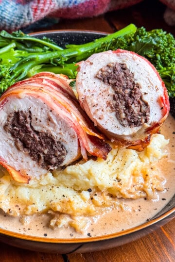 A plate with Chicken Balmoral: bacon-wrapped chicken stuffed with haggis, sliced to show the filling, served on creamy mashed potatoes with gravy, and a side of steamed broccolini.