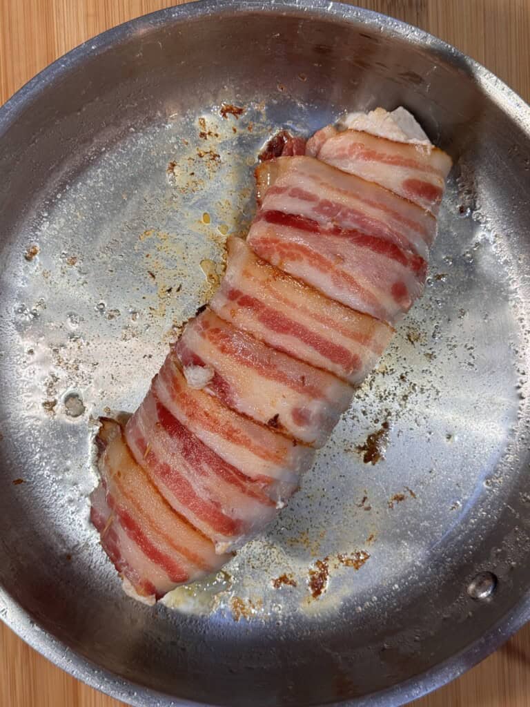 A seared Chicken Balmoral: bacon-wrapped meatloaf rests in a stainless steel pan, showing crispy, browned bacon strips on top. The pan has some rendered fat and browned bits around the meat. The surface beneath is wooden.