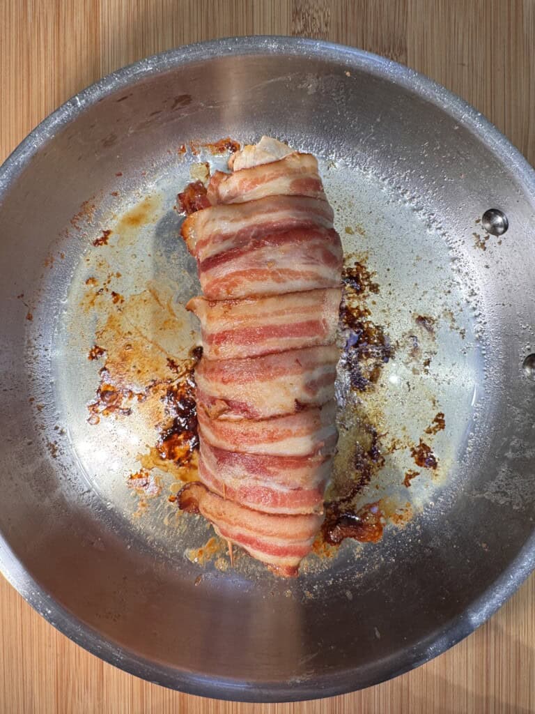 A cooked bacon-wrapped Chicken Balmoral sits in the center of a round metal pan on a wooden surface, with crispy bacon strips on top and browned juices around it.