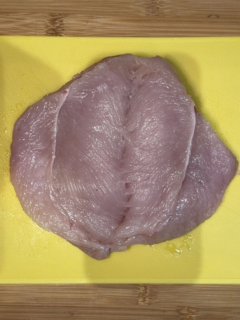 A raw, boneless chicken breast is spread flat on a yellow cutting board, placed on a wooden surface, ready for preparation.