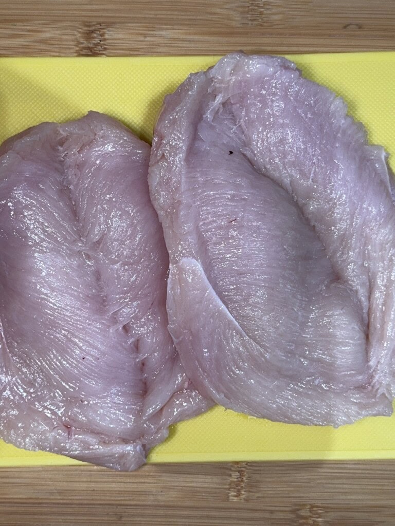 Two raw boneless, skinless chicken breasts are placed side by side on a yellow cutting board, which sits on a wooden surface.