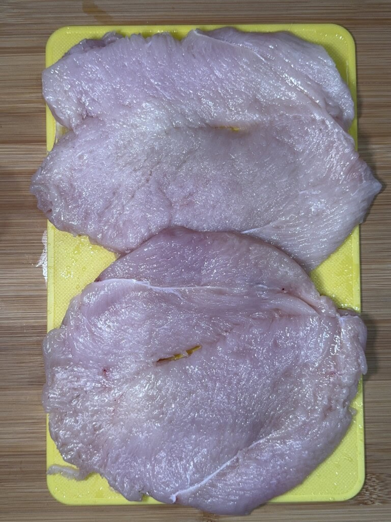 Two raw, thinly sliced pieces of chicken breast placed on a yellow plastic cutting board, set on a wooden surface.