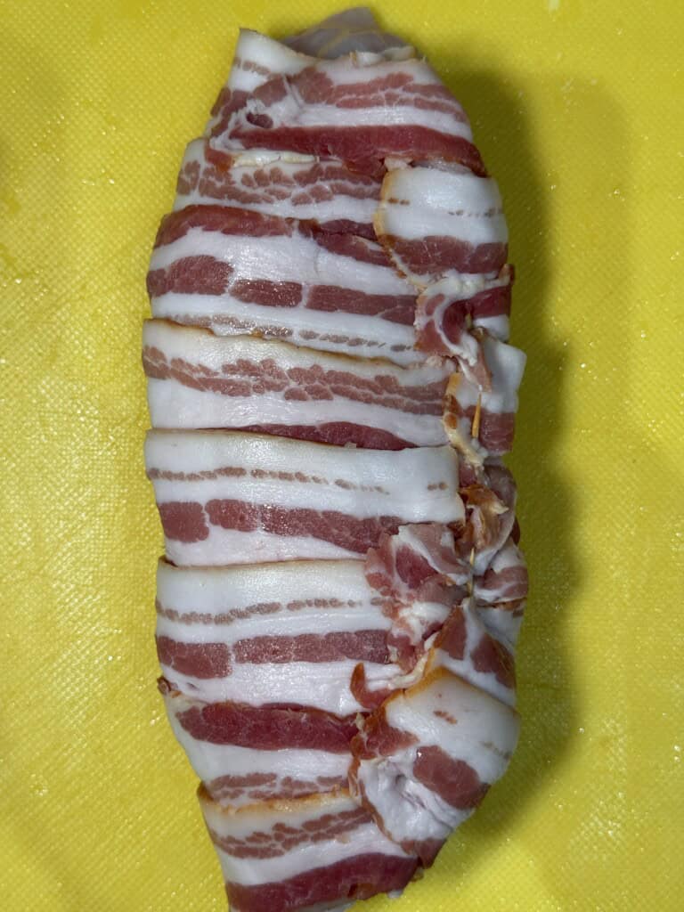 A raw, uncooked piece of chicken breast is wrapped in several strips of bacon and placed on a yellow cutting board.