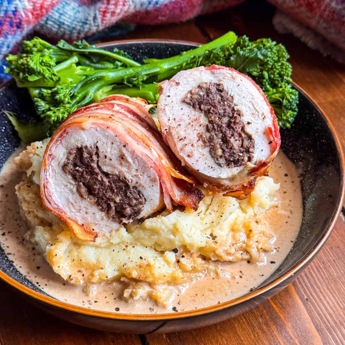 A plate with Chicken Balmoral: bacon-wrapped chicken stuffed with haggis, sliced to show the filling, served on creamy mashed potatoes with gravy, and a side of steamed broccolini.
