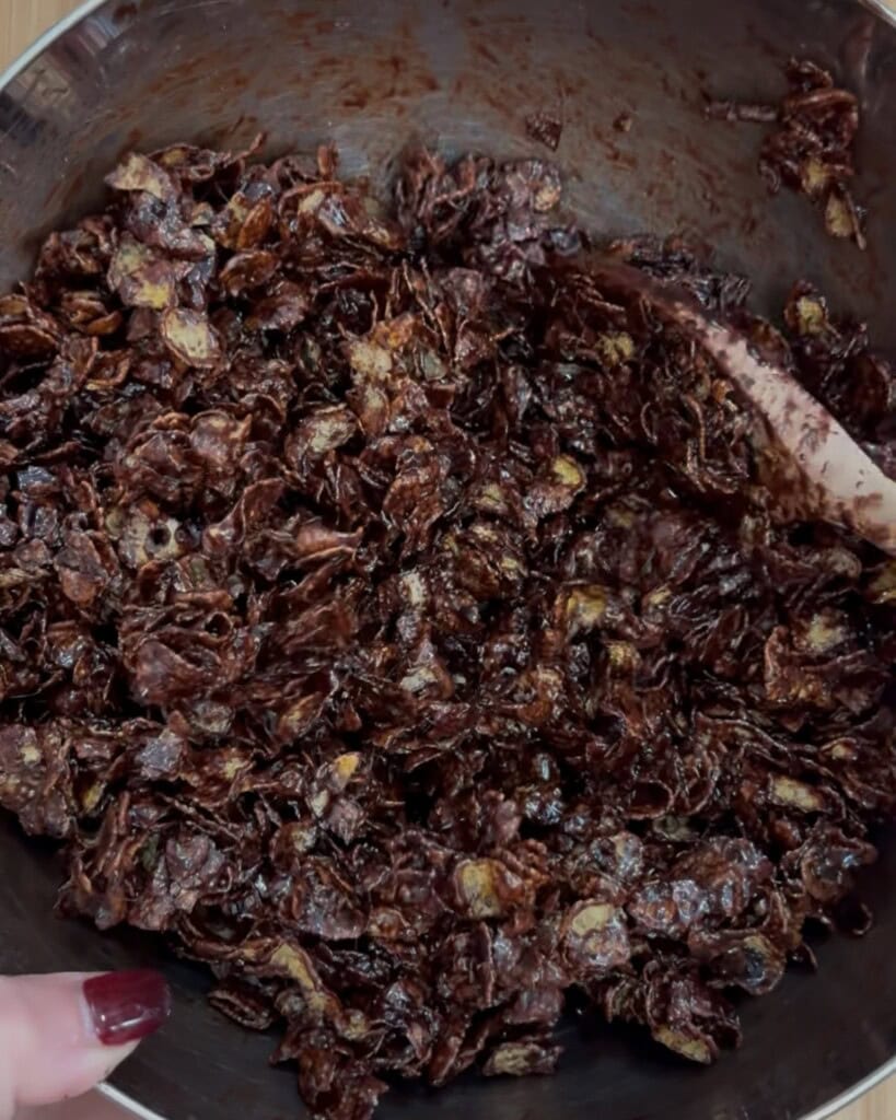 A close-up of a metal bowl filled with chocolate-covered cornflakes being mixed with a wooden spoon; a hand with red nail polish holds the bowl.