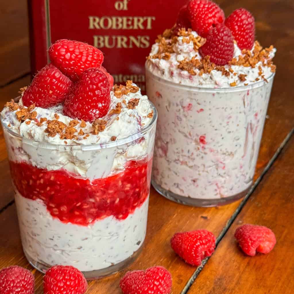 Two glass cups filled with creamy Cranachan, layered with raspberries and oats, and topped with fresh raspberries and crumbled topping, sit on a wooden table near a red book. Loose raspberries are scattered nearby.