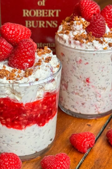 Two glass cups filled with creamy Cranachan, layered with raspberries and oats, and topped with fresh raspberries and crumbled topping, sit on a wooden table near a red book. Loose raspberries are scattered nearby.
