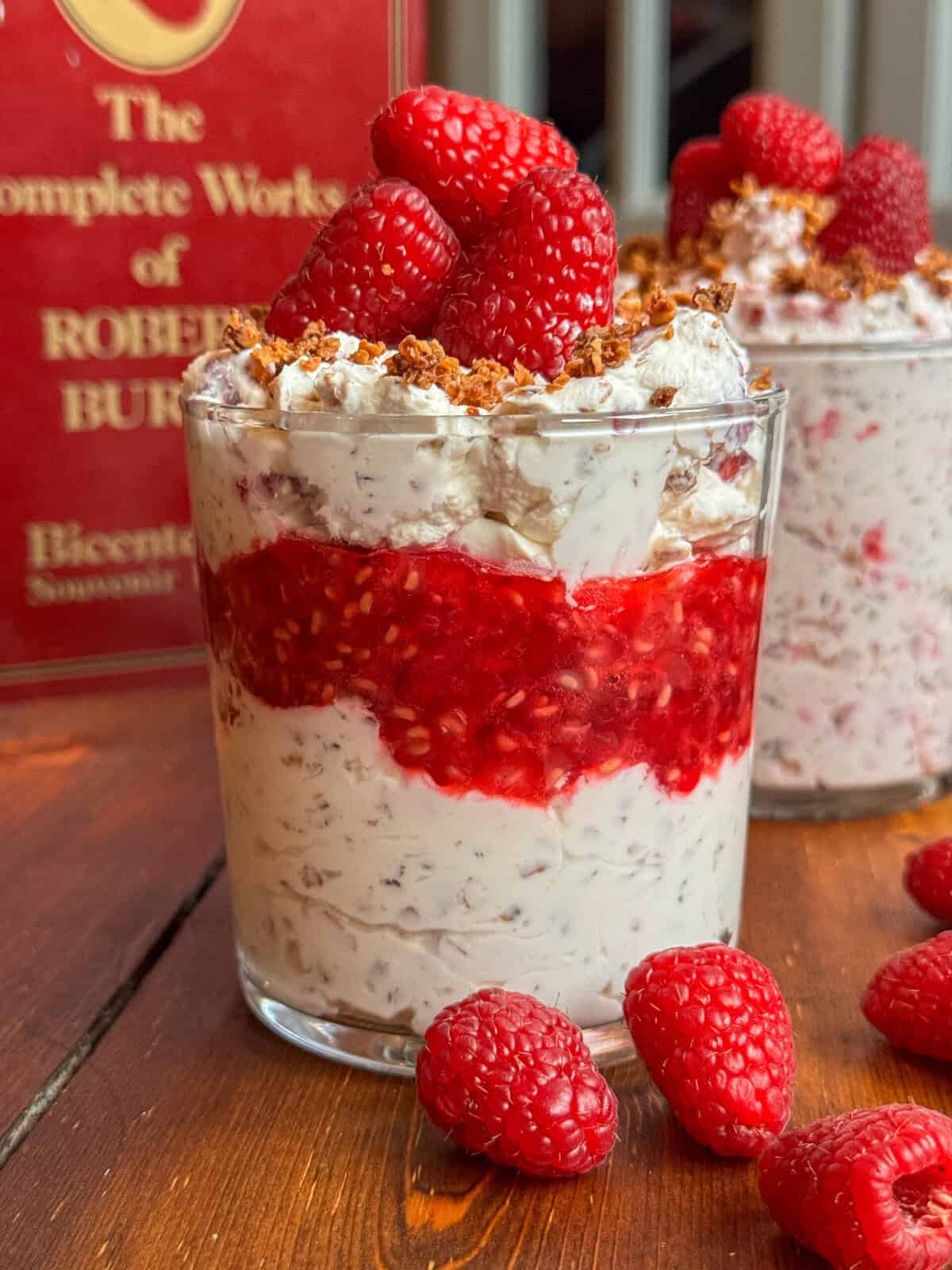 A glass filled with creamy Cranachan, layered with red raspberry sauce and topped with fresh raspberries and crunchy bits. Another similar glass and scattered raspberries are in the background, next to a red book.