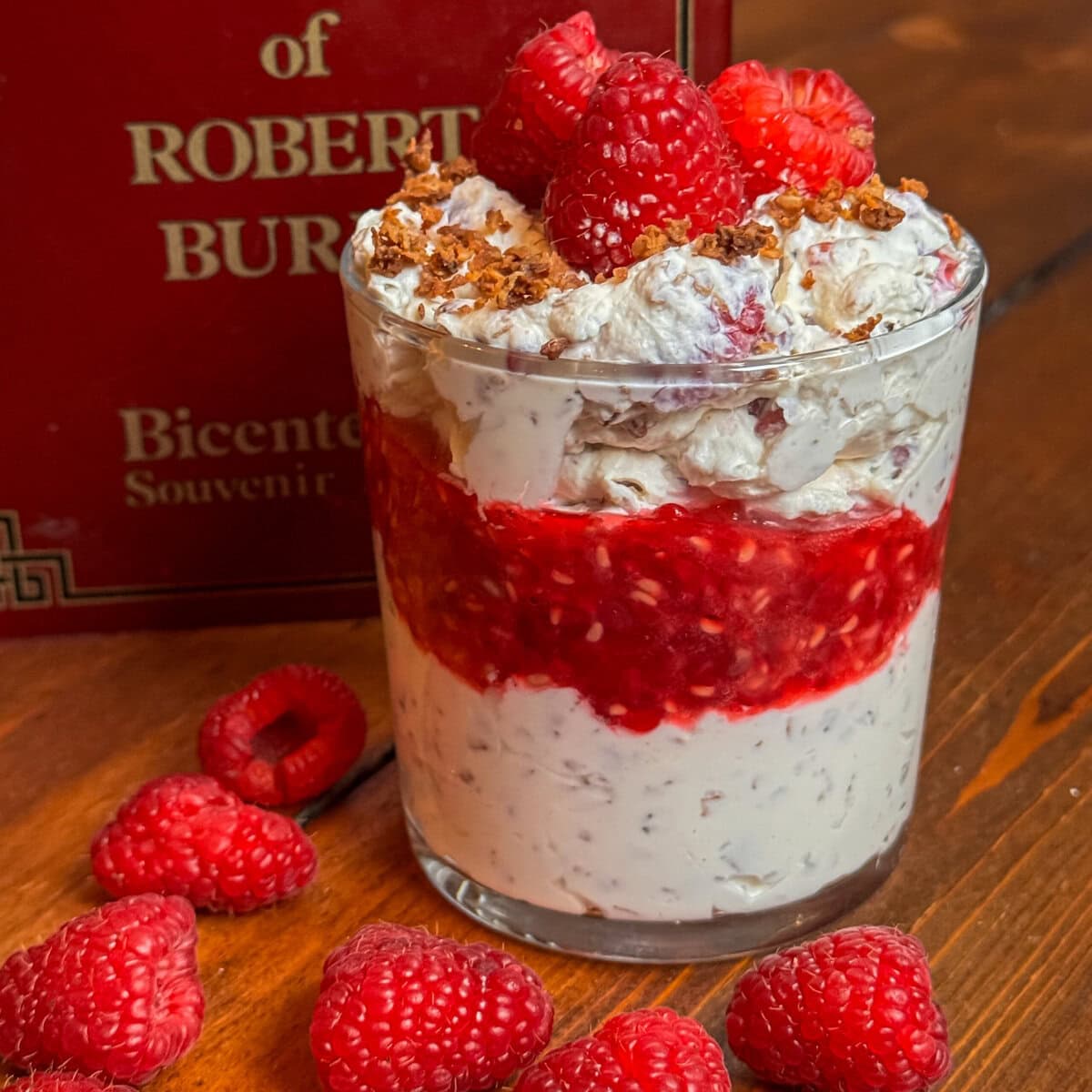 A glass filled with layers of creamy Cranachan, raspberry sauce, and topped with fresh raspberries and crunchy crumbs, sits on a wooden surface beside scattered raspberries and a red book.