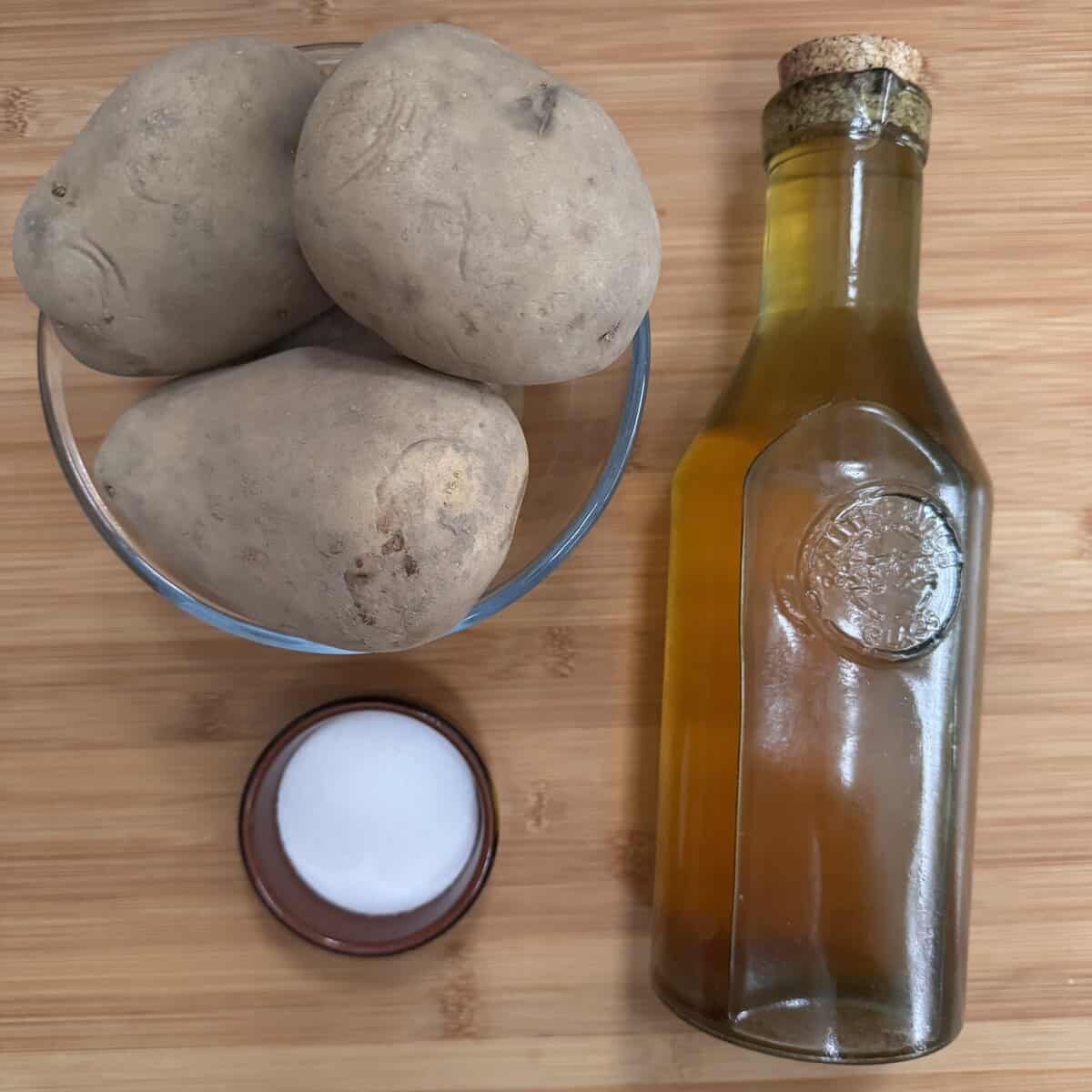 A glass bowl with large unpeeled potatoes, a round container of salt, and a bottle of vegetable oil with a cork top on a wooden surface.