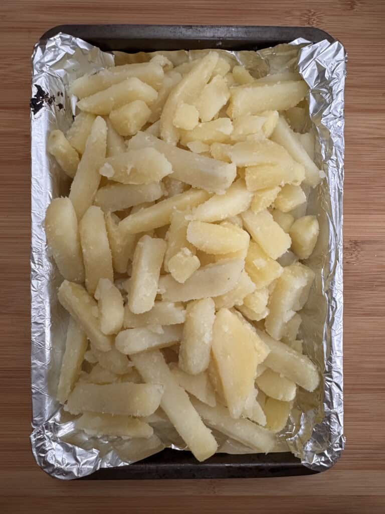 A metal baking tray lined with aluminum foil is filled with a pile of par-boiled homemade chips. The tray rests on a wooden surface.
