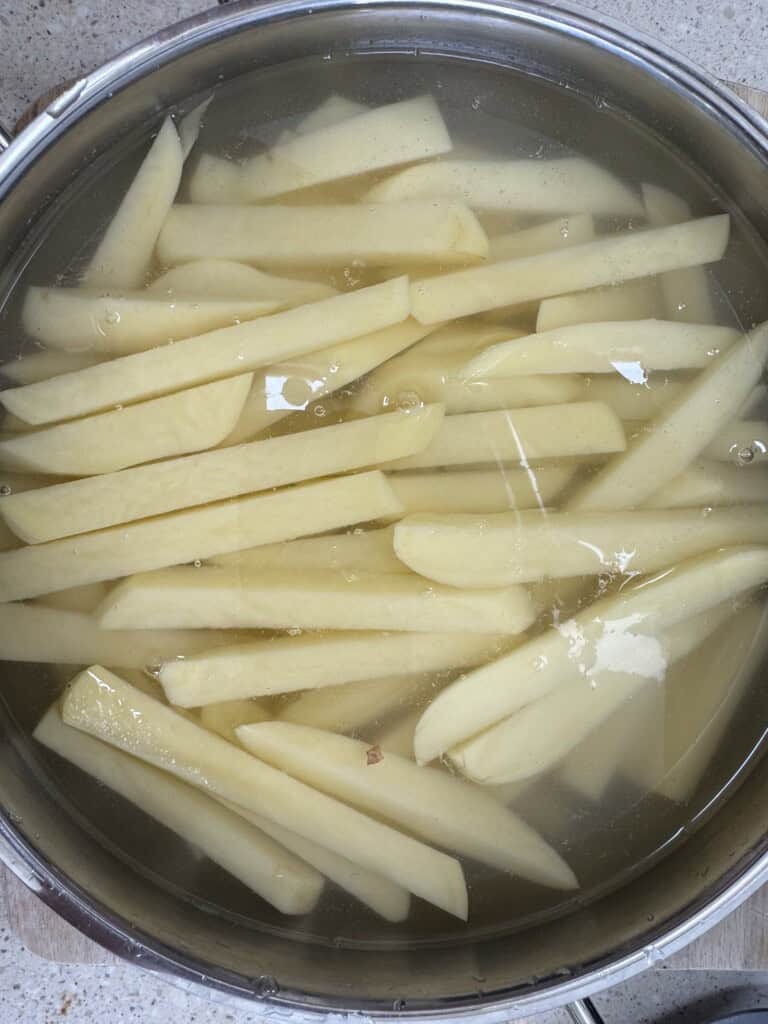 A metal bowl filled with raw potato slices soaking in water, likely being prepared for cooking. The potatoes are cut into thick strips, resembling French fry shapes.