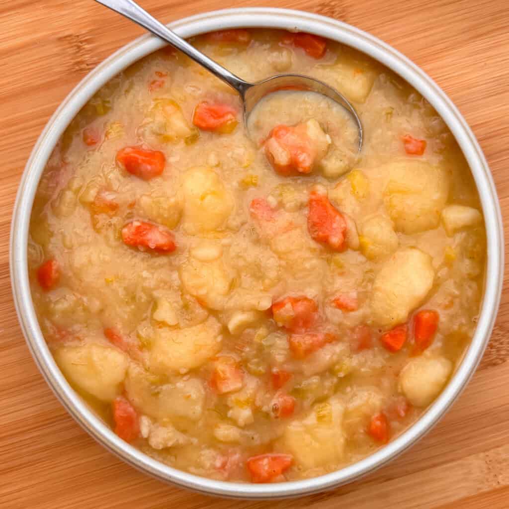 A bowl of Scottish tattie soup, featuring chunks of potatoes and carrots in a creamy broth, with a spoon resting inside. The bowl sits on a wooden surface.