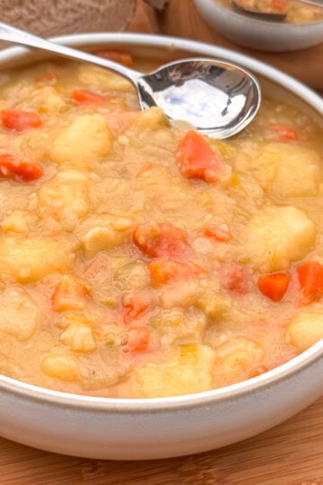 A bowl of Scottish tattie soup with chunks of potatoes and carrots, served with a metal spoon on a wooden table. Slices of bread are visible in the background.