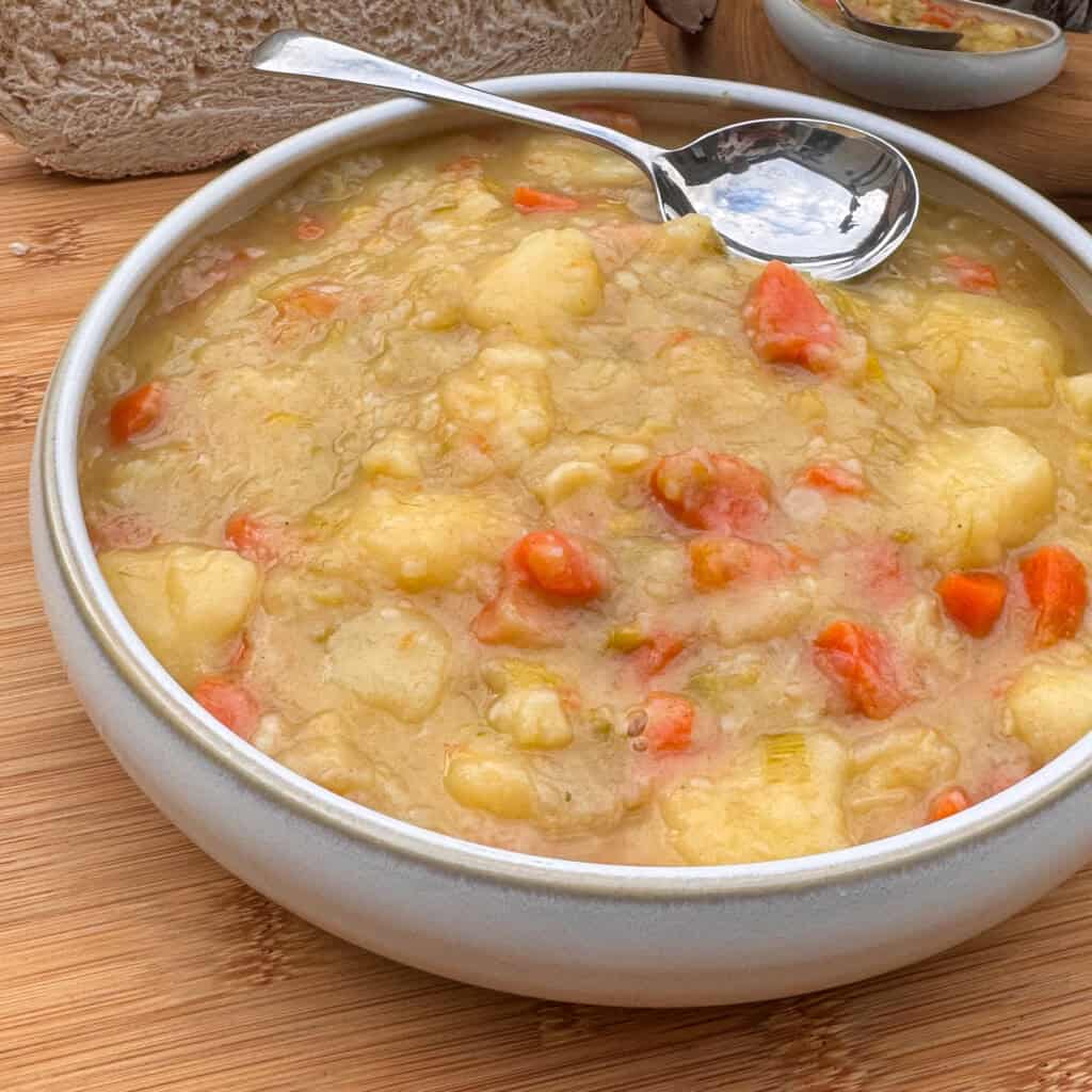 A bowl of Scottish tattie soup with chunks of potatoes and carrots, served with a spoon. Slices of bread and another small bowl are visible in the background on a wooden surface.