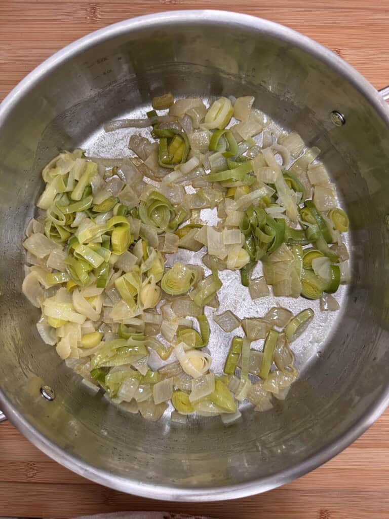 Chopped onions and leeks are being sautéed in a stainless steel pot on a wooden surface. The vegetables are lightly cooked and starting to soften, with a slight glisten from oil or butter.