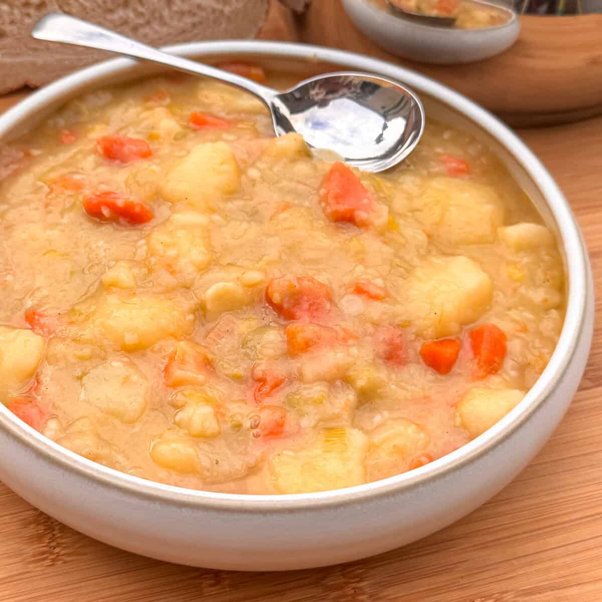 A bowl of Scottish tattie soup with chunks of potatoes and carrots, served with a metal spoon on a wooden table. Slices of bread are visible in the background.