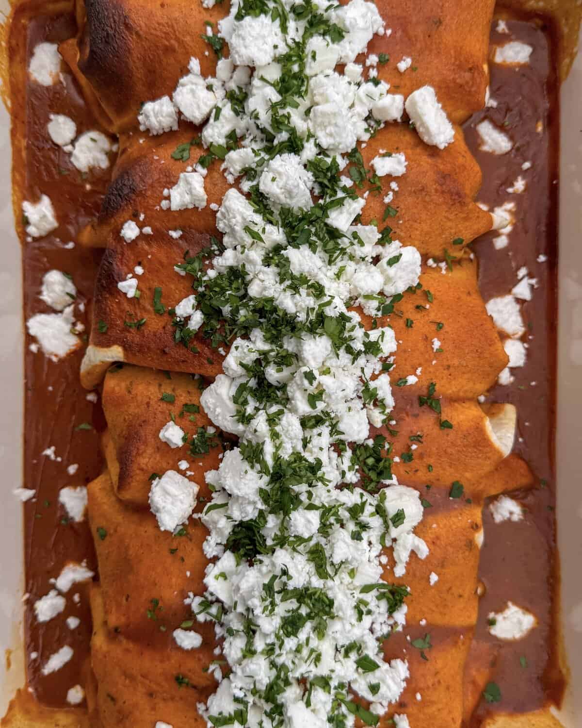 A close-up of baked enchiladas covered in red sauce, topped with a line of crumbled white cheese and chopped fresh herbs down the center.