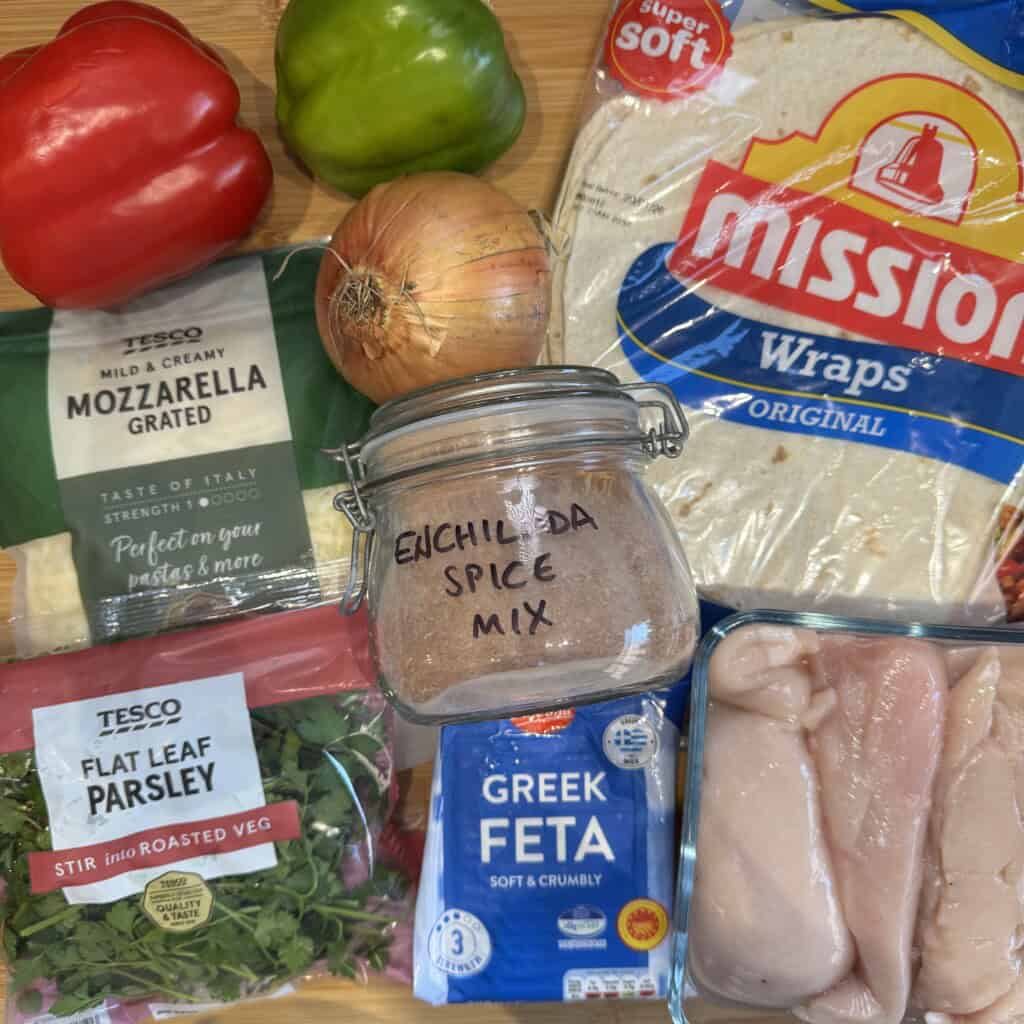 A selection of ingredients on a wooden surface: red and green bell peppers, original flour tortillas, grated mozzarella, a jar labeled enchilada spice mix, flat leaf parsley, Greek feta, an onion, and raw chicken breasts.