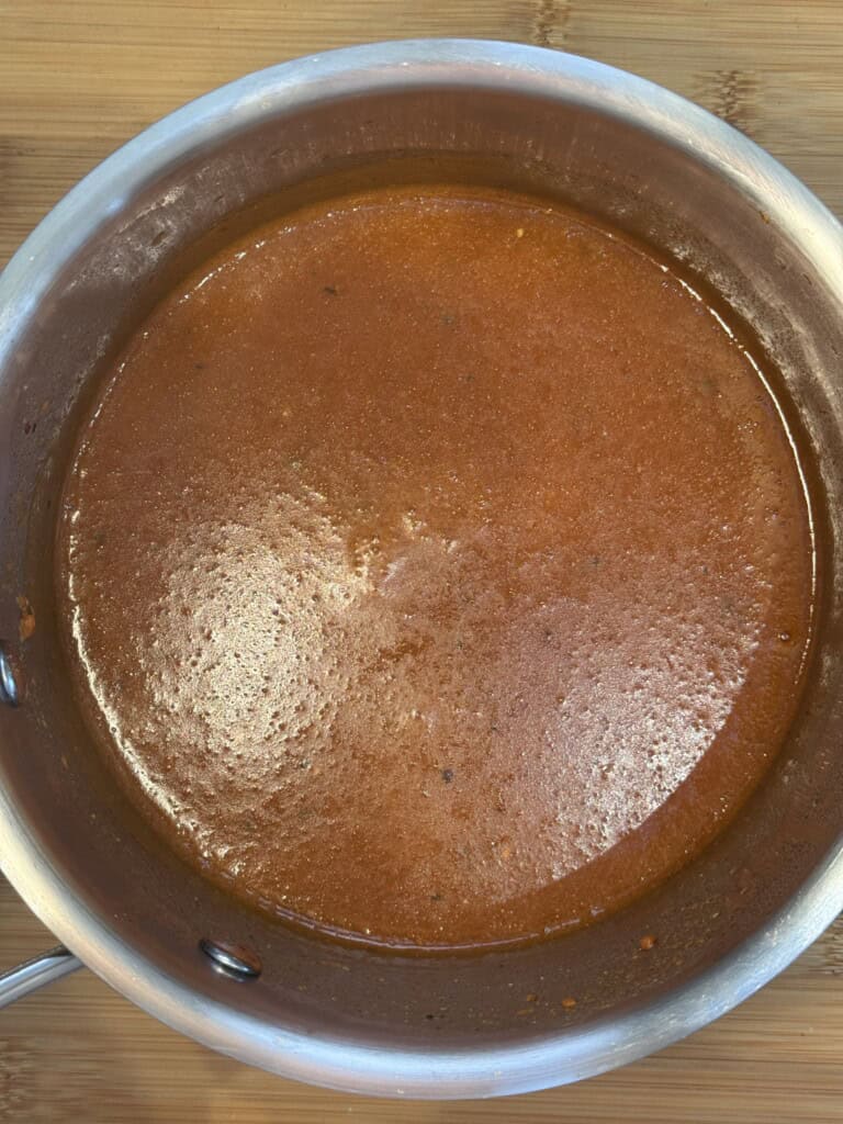 A stainless steel pot filled with smooth, red tomato sauce sits on a wooden surface. The sauce has a glossy texture and appears freshly cooked.