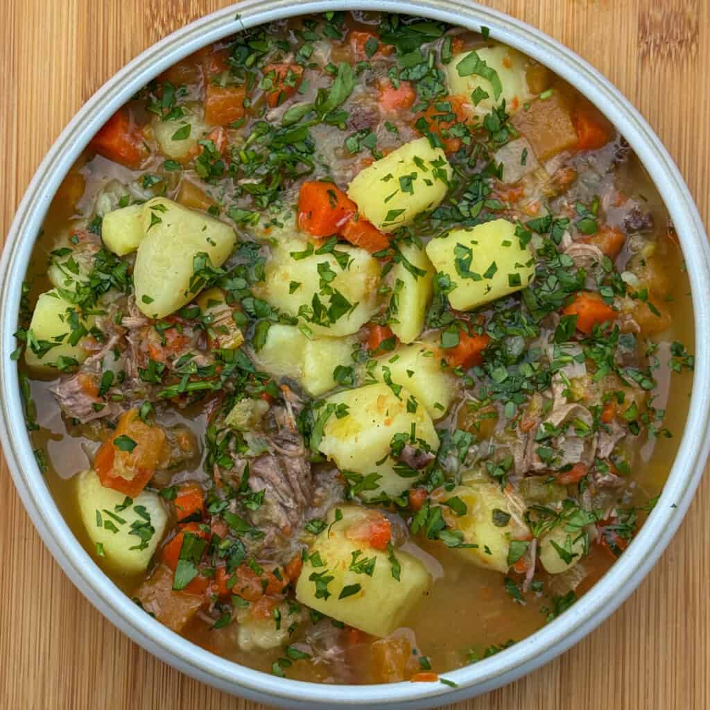 A bowl of hearty Welsh Cawl Soup with chunks of potato, carrots, and lamb, garnished with chopped fresh parsley, set on a wooden surface.