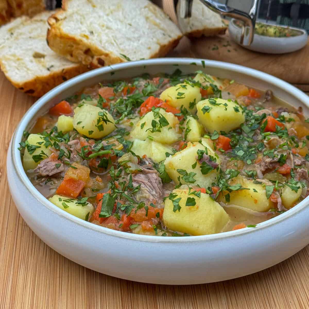A bowl of hearty Welsh Cawl Soup with chunks of potatoes, lamb, carrots, and parsley, served on a wooden surface with slices of bread in the background.