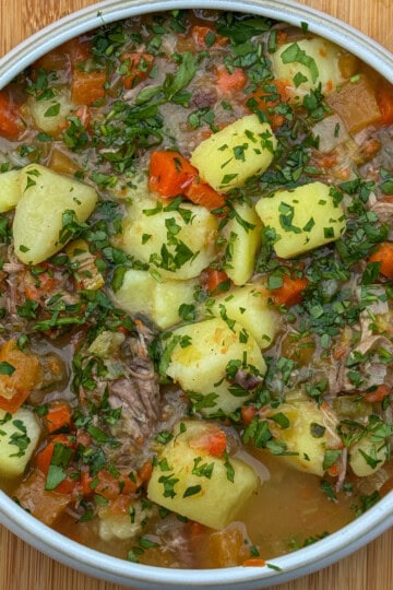 A bowl of hearty Welsh Cawl Soup with chunks of potato, carrots, and lamb, garnished with chopped fresh parsley, set on a wooden surface.