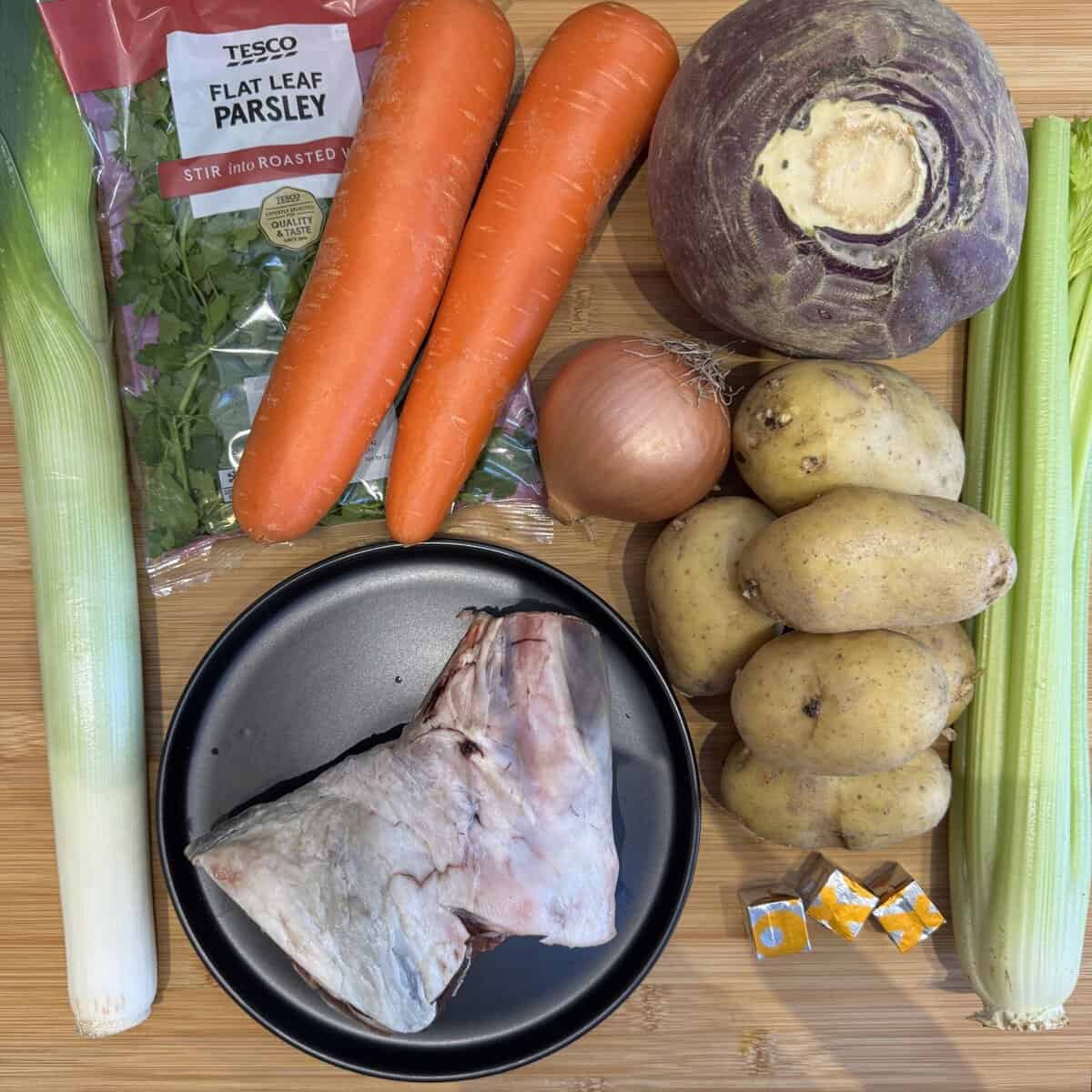 A leek, flat-leaf parsley, two carrots, a swede, an onion, four potatoes, two celery stalks, a lamb shank on a black plate, and two stock cubes arranged on a wooden surface.