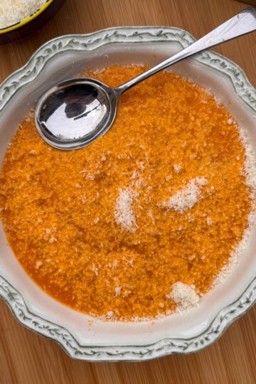 A bowl of Italian penicillin soup (pastina) topped with grated cheese, served in a decorative white bowl with a silver spoon resting on the edge, on a wooden surface.