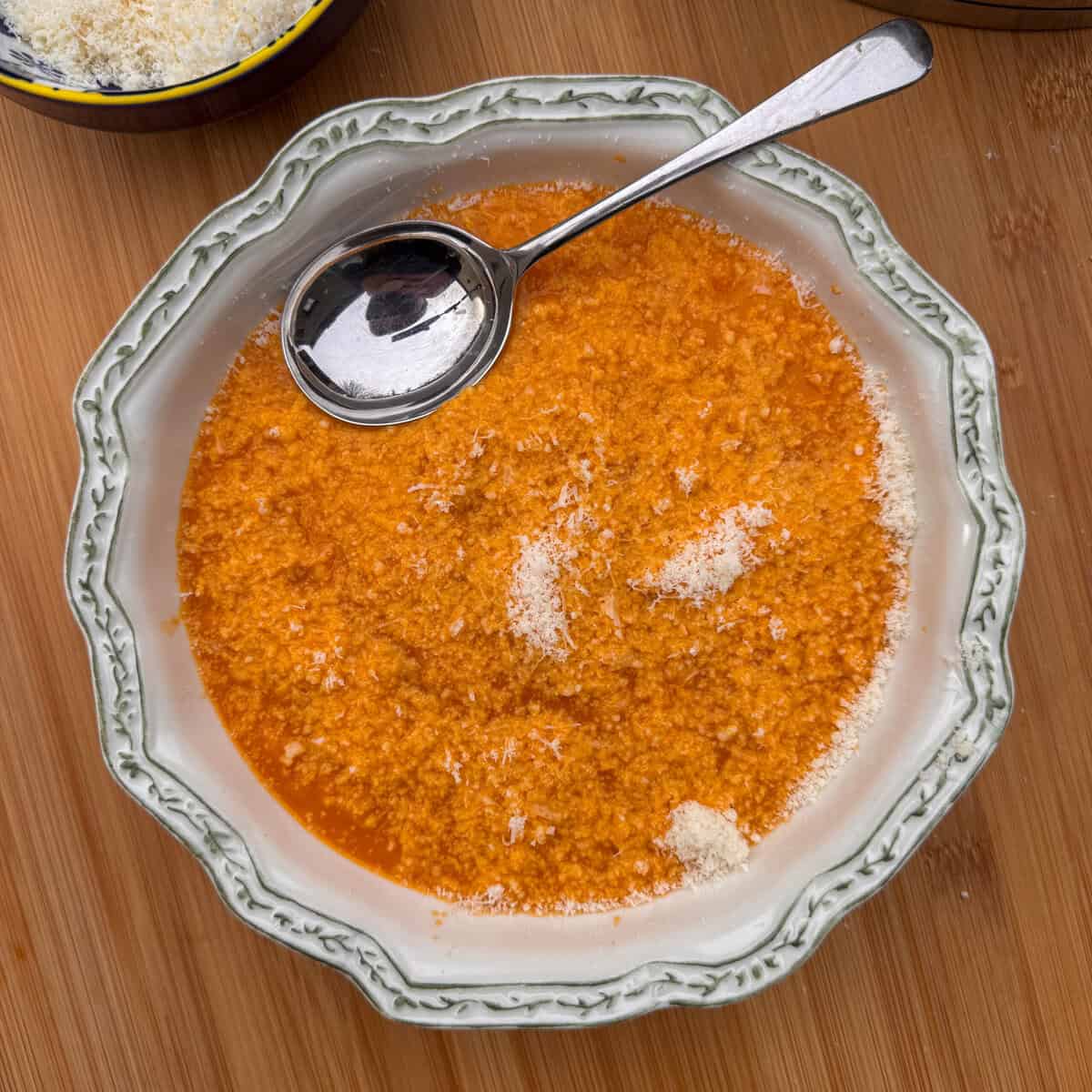 A bowl of Italian penicillin soup (pastina) topped with grated cheese, served in a decorative white bowl with a silver spoon resting on the edge, on a wooden surface.