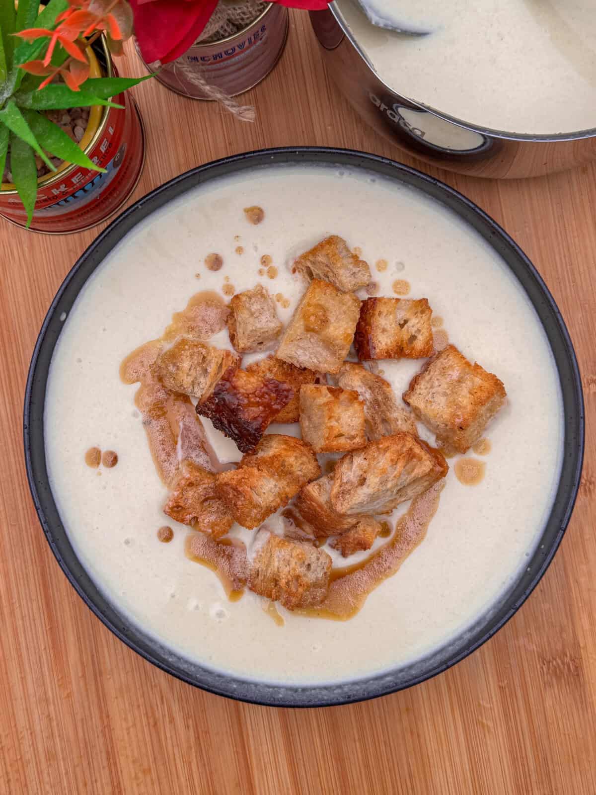 A bowl of cream of celery soup topped with golden-brown croutons and a drizzle of oil, set on a wooden surface with a potted plant and a pot of soup partially visible in the background.