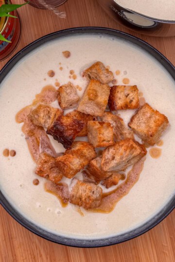 A bowl of cream of celery soup topped with golden-brown croutons, drizzled with oil, sits on a wooden surface with a potted plant and part of a pot visible in the background.