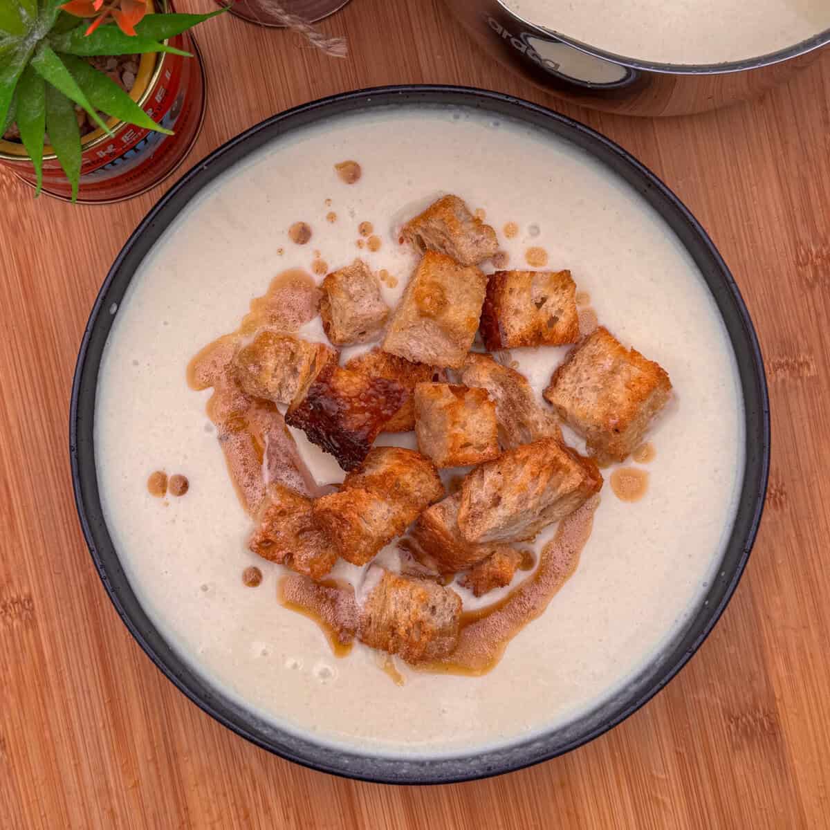 A bowl of cream of celery soup topped with golden-brown croutons, drizzled with oil, sits on a wooden surface with a potted plant and part of a pot visible in the background.