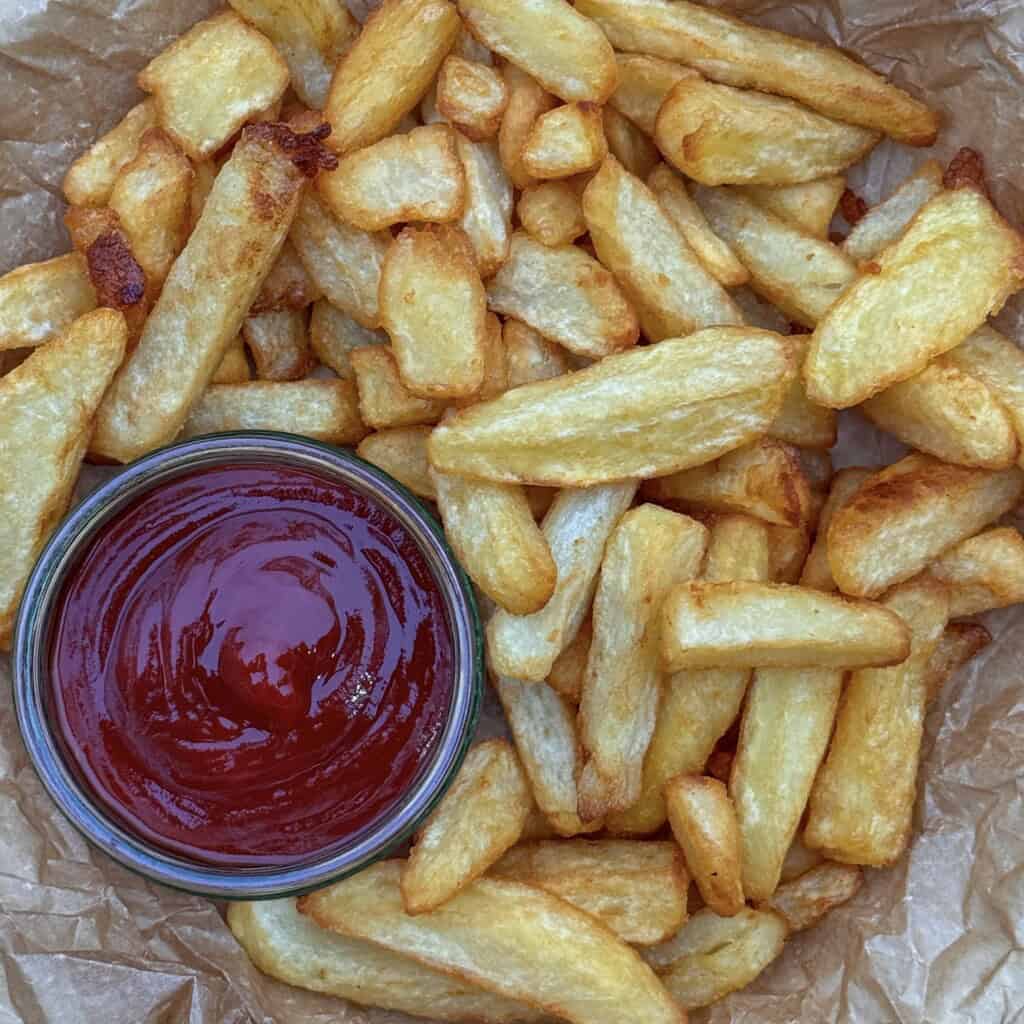 A pile of golden, crispy homemade deep fried chips on parchment paper, with a glass bowl of ketchup placed to the side for dipping.